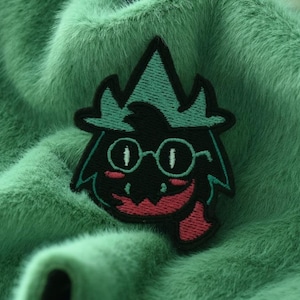 以下が含まれることがあります： 緑、黒、赤の刺繍パッチ。帽子と眼鏡をかけたキャラクターが大きく歯を見せて笑っている。