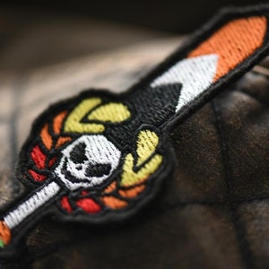 Könnte beinhalten: Ein schwarzer gestickter Aufnäher mit einem Totenkopf- und Knochenkreuz-Motiv. Der Totenkopf ist weiß mit schwarzen Augen und die Knochenkreuze sind orange, gelb und rot. Der Aufnäher ist auf einem braunen Stoff befestigt.
