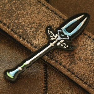 May include: An embroidered patch depicting a stylised sword with a white handle and a blue blade. The patch is blue and white and is on a brown leather background.