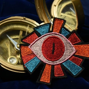 May include: A colorful embroidered eye patch with a red eye and black pupil, surrounded by orange, pink, and blue geometric shapes. The patch is on top of a brass compass.