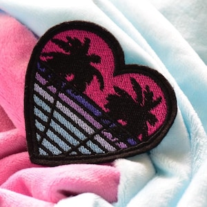 May include: A black heart-shaped patch with a pink and blue sunset scene and palm trees. The patch is on a pink and blue fabric background.