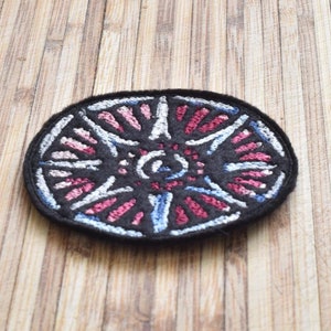 May include: A black embroidered patch with a white, pink and blue circular design. The design features a central circle with radiating lines.