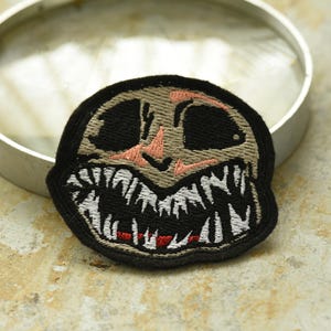 May include: Embroidered patch featuring a cartoon-style face with a wide, toothy grin. The face is beige with black eyes and a pink nose. The teeth are white with a red outline.
