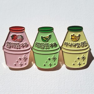 May include: Three enamel pins shaped like milk cartons with different fruit flavors. The pink carton has a strawberry and says "딸기맛 우유" in Korean. The green carton has a banana and says "바나나맛 우유" in Korean. The yellow carton has a melon and says "메론맛 우유" in Korean.