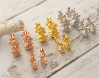 May include: Several pairs of floral hoop earrings in rose gold, gold, and silver tones are displayed. Each earring features a delicate flower design with small, sparkling accents. The earrings are arranged on a light-colored, wood-grain surface.