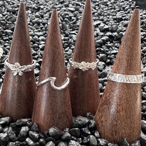 May include: Four silver rings displayed on wooden cone-shaped stands. The rings feature various designs, including a flower, a wave, and a band with the word "HAWAII". The rings are set against a backdrop of black pebbles.
