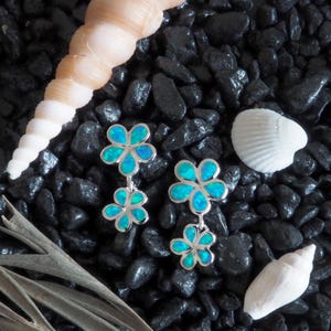 May include: A pair of silver earrings with a flower design, each flower having a blue and green opal center. The earrings are displayed on a bed of black stones, with seashells in the background.