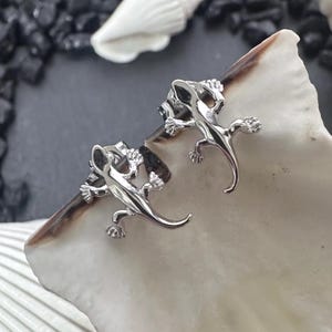 May include: A pair of silver-toned stud earrings shaped like small lizards. Each earring features a detailed gecko design with a smooth, reflective metallic finish, splayed legs, and a curved tail, secured with a standard post and butterfly back.