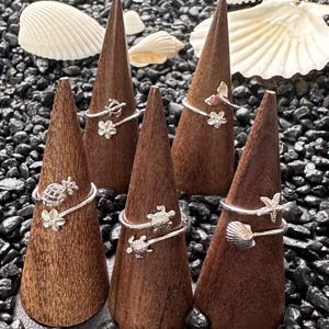 May include: Silver rings with ocean-themed designs, including a pineapple, turtles, starfish, and seashells, displayed on wooden cones. The rings are showcased against a backdrop of black pebbles and seashells.
