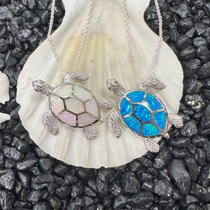 May include: Two silver turtle pendant necklaces. One turtle pendant has a white and iridescent shell, the other has a blue shell. The necklaces are displayed on a white seashell, surrounded by black stones.