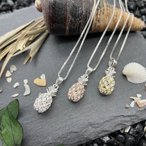 May include: Three silver necklaces with pineapple pendants. The pendants are in silver, rose gold, and gold. The necklaces are displayed on a dark gray slate surface with decorative seashells and small stones.