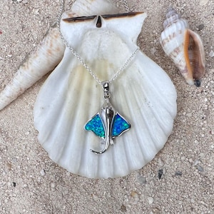 May include: A silver-toned necklace featuring a pendant shaped like a manta ray. The pendant has a blue and green opal-like inlay, with a detailed design. The necklace is displayed on a white seashell, with sand and other seashells in the background.