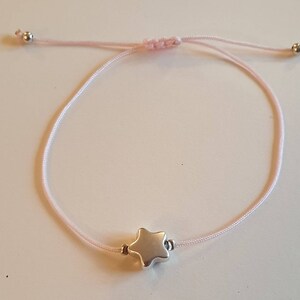 Könnte beinhalten: Ein hellrosa verstellbares Armband mit einem silbernen Sternanhänger. Das Armband besteht aus einer dünnen, gewebten Kordel und hat kleine silberne Perlen an den Enden. Der Sternanhänger ist der Mittelpunkt des Armbands.