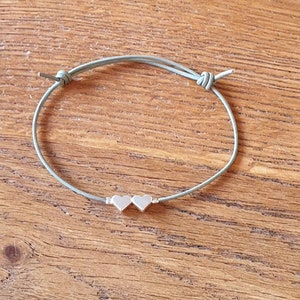 Könnte beinhalten: Ein hellgrünes Armband aus Kordel mit zwei silbernen Herzanhängern.