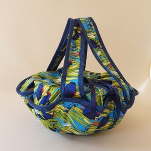 Puede incluir: Un bolso tote redondo de tela con un estampado de pájaros azules, verdes y amarillos. El bolso tiene dos asas azules y un ribete azul. El bolso está recogido en la parte superior con una cinta azul.