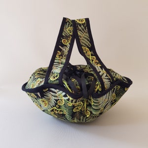 Puede incluir: Cesta de tela hecha a mano con un estampado de helechos verdes y amarillos sobre fondo negro. La cesta tiene dos asas negras y un ribete negro. El interior también es negro. Diseñada para almacenamiento decorativo.