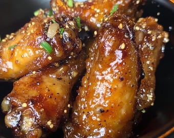 May include: Close-up of several chicken wings glazed with a dark sauce and sprinkled with sesame seeds. The wings are arranged in a bowl.