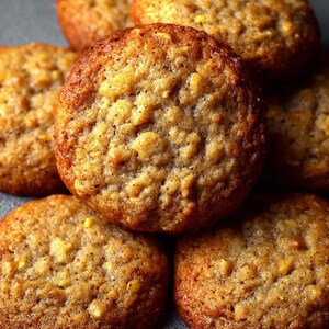 Puede incluir: Primer plano de varias galletas de color marrón dorado. Las galletas tienen una superficie texturizada con avena visible y un exterior ligeramente más oscuro y crujiente. Están apiladas y dispuestas, mostrando su forma redonda y su aspecto horneado.