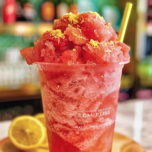 May include: A close-up of a red watermelon slushie with lemon zest in a clear plastic cup with a yellow straw. The cup has the text "REM PLISS" and "MALLCIMBA.COM" printed on it.