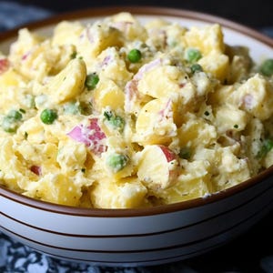 Può includere: Primo piano di una ciotola piena di insalata di patate. L'insalata presenta pezzi di patate, cipolla rossa e piselli verdi, il tutto mescolato in un condimento cremoso. La ciotola ha un interno bianco e un bordo a righe marroni e bianche.