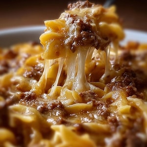 Puede incluir: Primer plano de una porción de pasta horneada con queso derretido y carne picada. El queso se estira al levantar la pasta, mostrando una comida cálida y acogedora. El plato es de un color dorado intenso, con énfasis en las texturas.