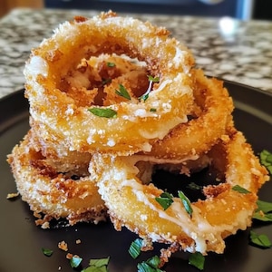 May include: A stack of golden brown onion rings, drizzled with a white sauce and garnished with fresh parsley, on a black plate.