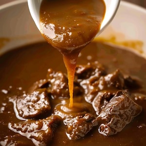 May include: A close-up shot shows brown gravy being poured from a white bowl onto pieces of meat. The gravy is thick and glossy, with a rich, dark colour. The meat looks well-cooked and tender. The image suggests a feeling of warmth and comfort, typical of a hearty meal.