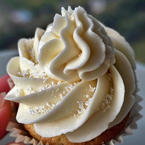 以下が含まれることがあります： オフホワイトのクリーム状のフロスティングが渦巻くカップケーキのクローズアップ。フロスティングは小さな白いスプリンクルで飾られています。カップケーキは白い紙のライナーに入っており、ケーキのベースは薄茶色です。