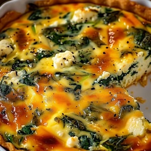 Pode incluir: Close-up de uma quiche com uma crosta dourada. O recheio é uma mistura de creme amarelo, espinafre verde e queijo branco. Pimenta preta é polvilhada por cima. Uma fatia foi removida, revelando as camadas.