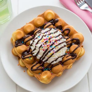 May include: A bubble waffle topped with whipped cream, chocolate syrup, and colorful sprinkles sits on a white plate. A glass of green liquid and silverware are in the background. The waffle has a unique, round, bubble-like texture.