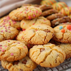 Può includere: Un mucchio di biscotti appena sfornati di colore marrone dorato. Alcuni biscotti sono decorati con zuccherini rosa e rossi, mentre altri hanno gocce di cioccolato e caramelle colorate. I biscotti sono disposti su una griglia di raffreddamento in metallo.