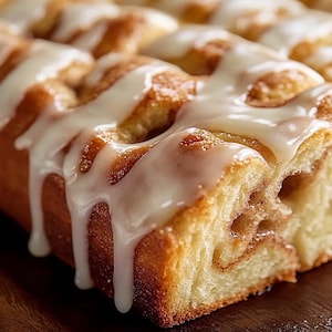 May include: Close-up of a cinnamon roll with a white glaze. The roll is cut into squares and has a swirl of cinnamon in the center. The glaze is dripping down the sides of the roll.