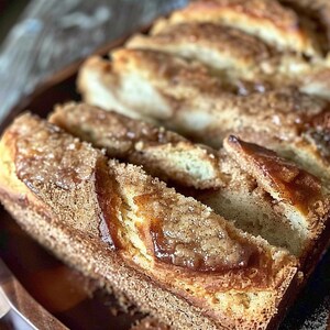 Puede incluir: Primer plano de una hogaza de pan de canela en rodajas. El pan tiene una corteza dorada con una generosa cobertura de azúcar de canela y un glaseado. El pan está sobre una bandeja de madera.