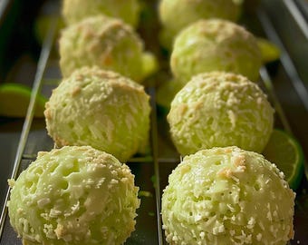 May include: Close-up of several lime-green, round desserts on a wire rack. The desserts are coated with a white, shredded topping. Slices of lime are scattered around the desserts, adding a fresh, vibrant touch. The desserts appear to be cooling.