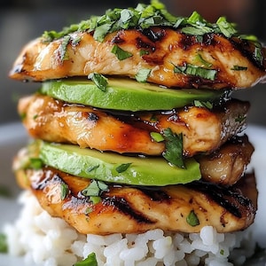 Puede incluir: Una pila de pechugas de pollo a la parrilla, intercaladas con rodajas de aguacate y adornadas con hierbas frescas, se asienta sobre una cama de arroz blanco. El pollo tiene marcas de parrilla y un glaseado brillante. El aguacate es verde brillante y las hierbas están finamente picadas.