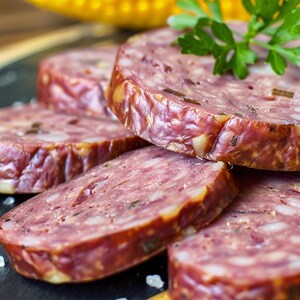 Può includere: Primo piano di salame a fette, con un esterno marrone rossiccio e un interno marmorizzato. Guarnito con prezzemolo fresco, il salame è disposto su una superficie scura, con cristalli di sale visibili. Una delizia culinaria.