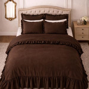 May include: A bed dressed with a brown ruffled duvet cover and matching pillowcases. The bed has a buttoned headboard and is set against a neutral wall. A gold-framed mirror hangs on the wall to the left.