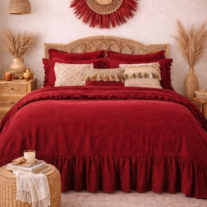 以下が含まれることがあります： 深い赤色の掛け布団と枕が置かれたベッドのある寝室のシーン。 ベッドには織りのヘッドボードがあり、装飾的なクッションが飾られています。 織りのオットマンと壁のアートがボヘミアンスタイルを演出しています。