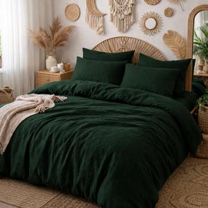 May include: A bedroom scene featuring a bed with dark green bedding, including a duvet cover and pillows. A beige throw blanket is draped over the bed. The room is decorated with woven wall hangings, a rattan headboard, and a woven pendant light, creating a bohemian aesthetic.