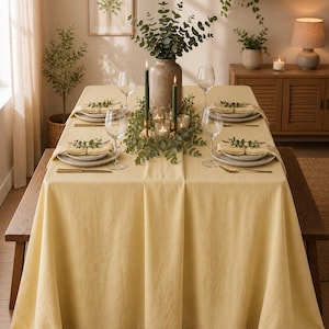 May include: A dining table set for a meal, featuring a pale yellow tablecloth. The table is set with plates, silverware, and wine glasses. A vase with greenery and candles are in the center, creating a warm and inviting atmosphere.