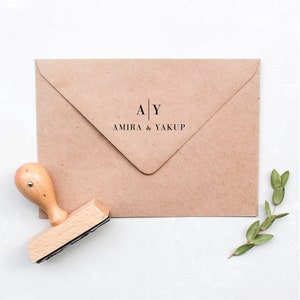 Könnte beinhalten: Ein brauner Kraftpapierumschlag mit dem Text "A | Y AMIRA & YAKUP" in Schwarz gedruckt. Ein Holzstempel mit hellem braunen Griff und schwarzem Stempelkissen liegt neben dem Umschlag. Ein Zweig mit grünen Blättern befindet sich in der unteren rechten Ecke.
