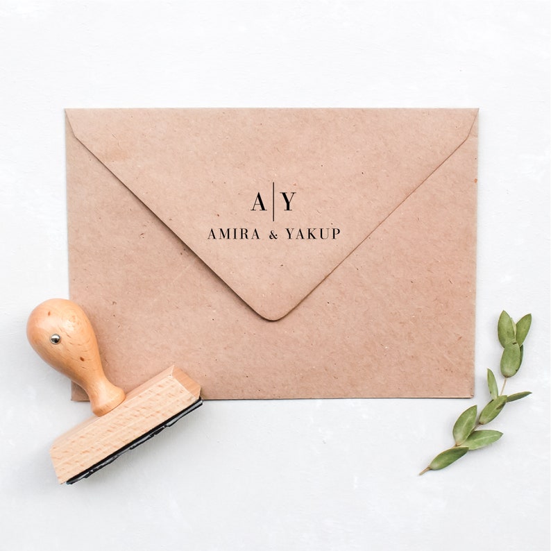May include: A kraft paper envelope with the text "A | Y AMIRA & YAKUP" printed in black. A wooden stamp with a light brown handle and a black ink pad is next to the envelope. A sprig of green leaves is in the lower right corner.
