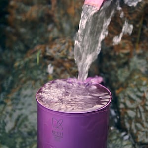 以下が含まれることがあります： 紫色の容器に、お揃いの紫色のピッチャーから注がれた水が入っています。容器には「KEITH TITANIUM」の文字が印刷されています。背景は岩と水のある自然な環境です。