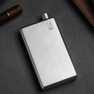 May include: A rectangular, silver-coloured flask with a small, rounded cap. The flask has the text "KEITH TITANIUM" printed on the front. The flask is on a dark grey surface, with a wooden object and a brown leather case in the background.