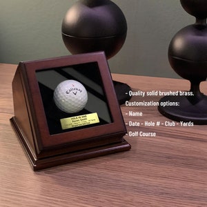 May include: A wooden display case with a glass front, showcasing a white golf ball with the Callaway logo. The case has a brass plaque with the text "HOLE IN ONE Raven Smith 1.12.2017 Hole 17 - 1 Iron - 120 Yards Winged Foot Golf Club".