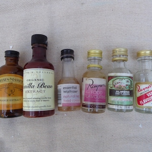 May include: A collection of six glass bottles containing different food flavorings. The bottles are labeled with the flavoring type, such as vanilla bean extract, almond food flavoring, and coffee food flavoring.