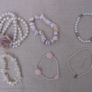 May include: An assortment of bracelets in various styles. One bracelet is made of white pearls and features a large pink rose. Other bracelets include shell, pearl, and bead designs. The bracelets are displayed on a neutral-colored surface.