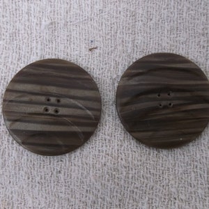Puede incluir: Dos botones grandes y redondos con aspecto de veta de madera marrón oscuro. Cada botón tiene cuatro agujeros para coser. Los botones están sobre un fondo de tela blanca texturizada.