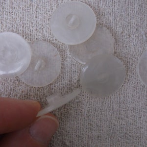 May include: A collection of translucent, round buttons with a pearlescent sheen. The buttons vary in size and have a two-hole design on the back. One button is held by a hand, showing the button's attachment piece. The buttons are on a textured, off-white surface.
