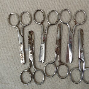 May include: A set of seven vintage silver-toned scissors with slightly rusted blades. The scissors are arranged in a row on a beige fabric background.